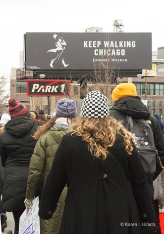 Marchers walking down Chicago street.  Above is a Johnny Walker billboard saying Keep Walking Chicago
